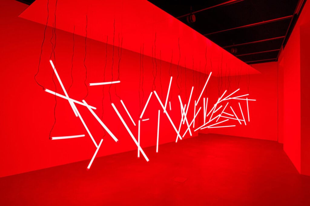 A room illuminated in red light features numerous white fluorescent tubes hanging and intersecting at various angles along the wall, creating a modern, abstract light installation for the Atto 2/3: Col Tempo exhibition.