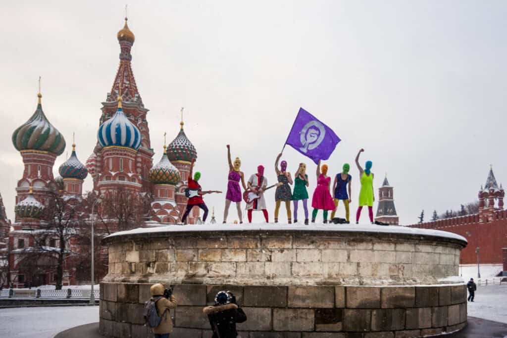 Maria Alyokhina of Pussy Riot and others, dressed in colorful outfits and masks, stand on a stone platform in Red Square, Moscow, holding a purple flag. St. Basil's Cathedral and the Kremlin loom behind them in the snow as two photographers capture the scene below.