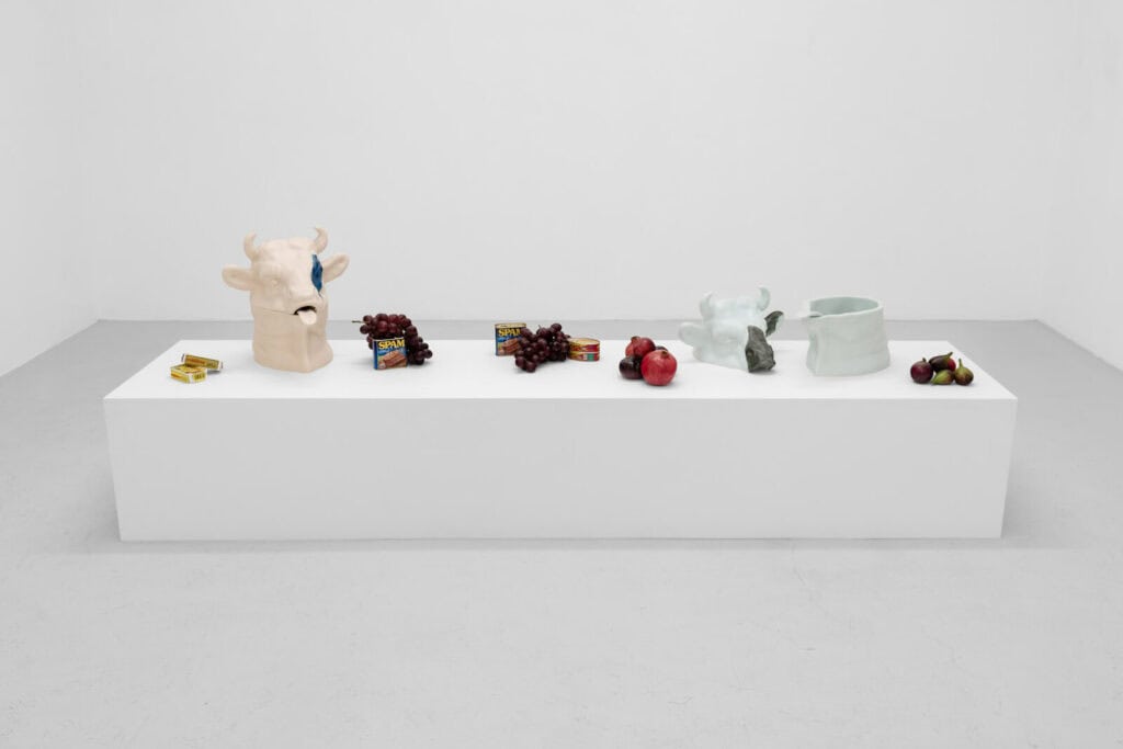 A white rectangular pedestal displays two ceramic cow heads, fruit including grapes and apples, three Spam cans, a light green ceramic vessel, and a small pile of plums in a minimalist white gallery space evoking the playful spirit of Sailor Moon ‘Abigail’s Party’ at Rose Easton Gallery.