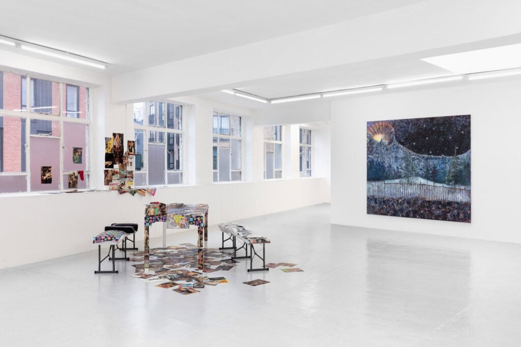 A bright, white-walled art gallery with large windows displays a colorful night scene painting. Near the windows, a table and benches are scattered with photos and papers—setting the stage for ‘Abigail’s Party’ at Rose Easton Gallery.