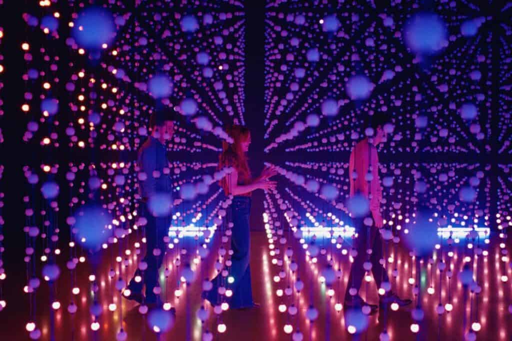 Three people stand among hanging strings of glowing orbs in a dark room, illuminated by purple and blue lights, creating an immersive, futuristic, and surreal atmosphere.