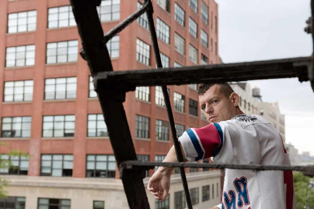A man in a white and red sports jersey leaning against a black metal fire escape structure on a New York rooftop, gazing sideways against an overcast sky — photograph by Slava Mogutin
