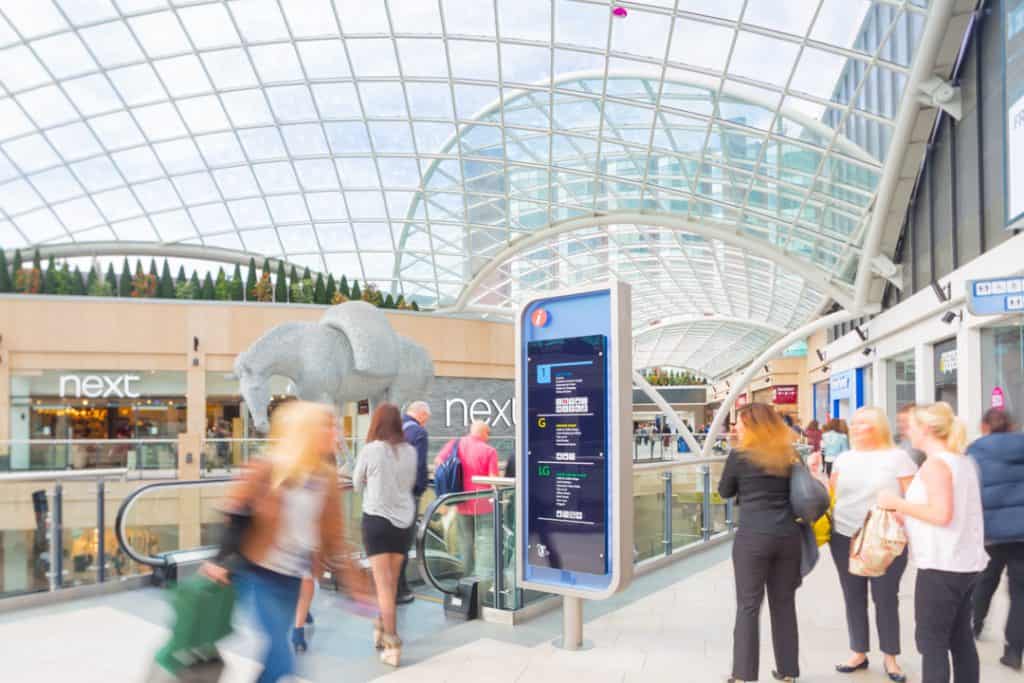Lozenge shaped index sign inset in blue acrylic located at the top of the escalator within Trinity Leeds Shopping Centre, with shopper passing by. In the background is a large metal sculpture of a horse and the glass panelled, curved roof