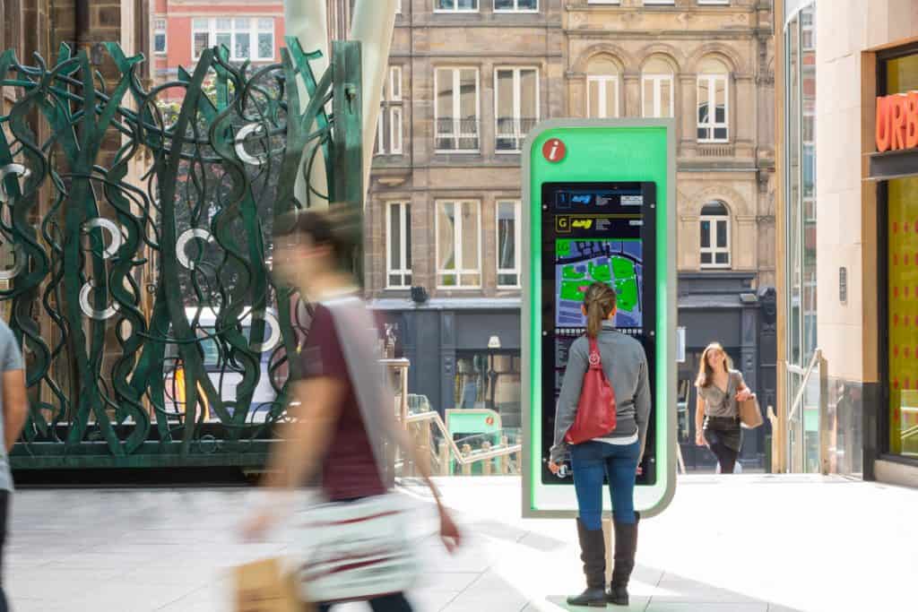 Image of a totem sign, located on a ground floor entrance to the Trinity Leeds Shopping Centre. The sign is a vertically oriented lozenge shape with a transparent green acrylic infill. The information is displayed on a black rectangular panel and includes a plan of the shops and a retailer list.
