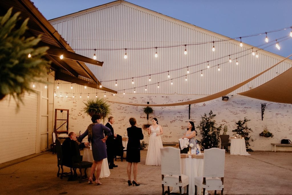Outdoor Patio space on a summer evening. The patio space features string lights and sun shades, along with white washed brick and ferns/greenery. A Bride speaking with some of her guests are at the center of the image.