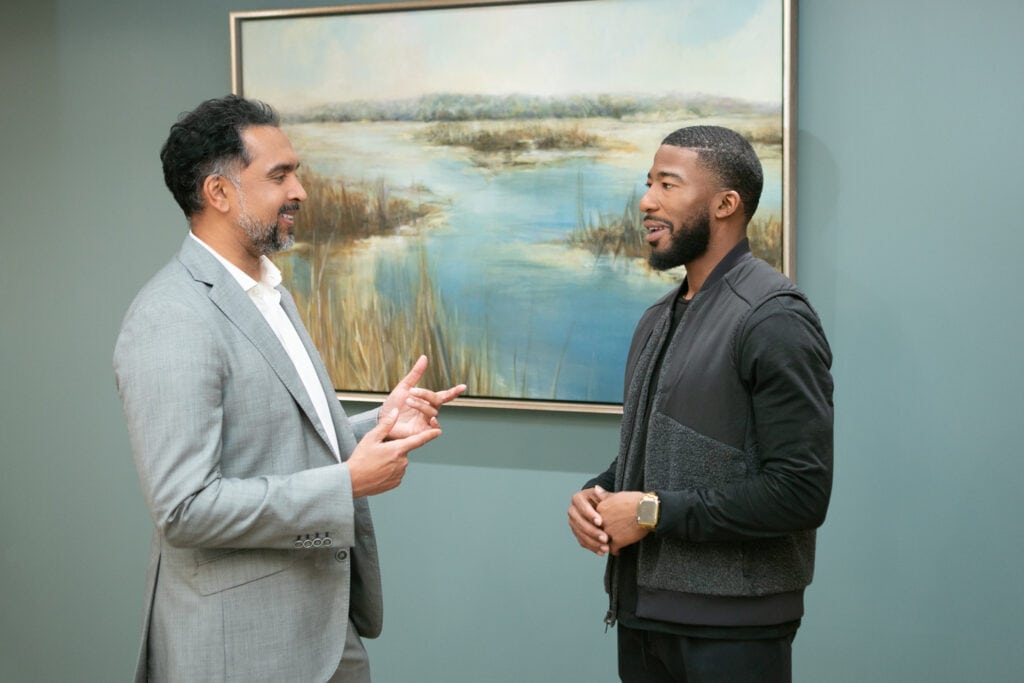 Two men stand and talk in front of a landscape painting; one, Dr. Murthy, wears a gray suit, while the other sports a dark casual outfit.