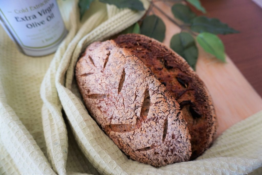 Olive sourdough with wheat pattern scoring on a cutting board with green towel, fake leaves and an olive oil can