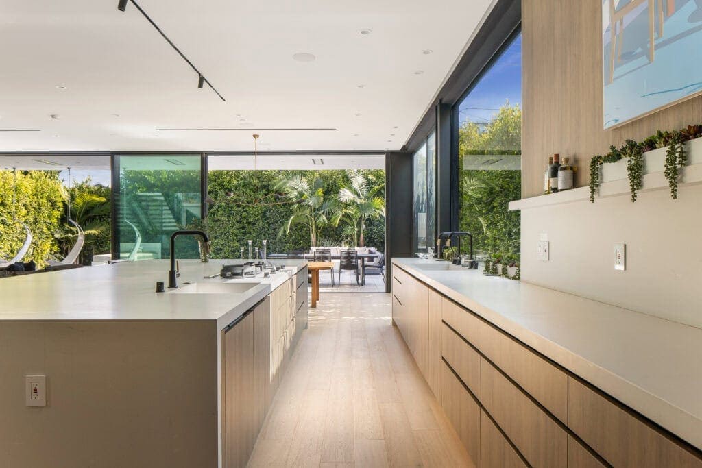 Contemporary kitchen with minimalist design, white surfaces, natural wood cabinetry, expansive glass sliding doors showing lush greenery outside.