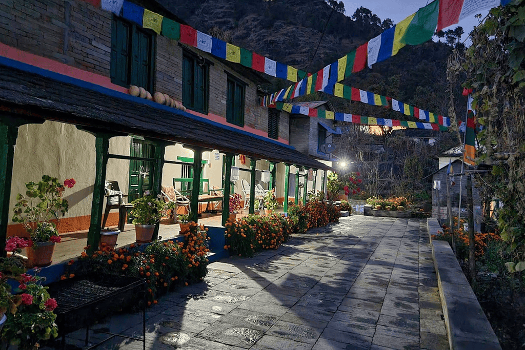 A traditional two-story rural Nepalese homestay featuring brick walls, wooden pillars, potted marigold flowers along the porch, and colorful Buddhist prayer flags strung across the yard.