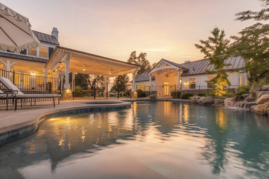 3itOT-gigapixel-low resolution v2-4x • Jaydee Sheppard Real Estate Poolside view of a resort at sunset with white buildings, string lights, and lounge chairs by a curved pool.