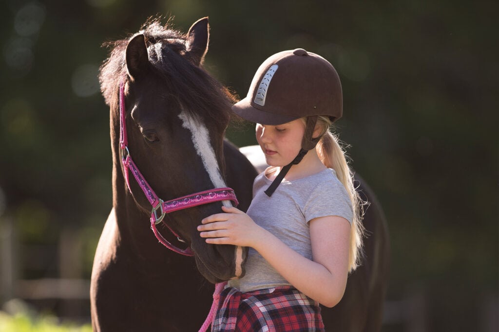 Flicka med ridhjälm klappar en svart häst med vit bläs i ansiktet, båda står i solljus på en sommargrön ridanläggning.