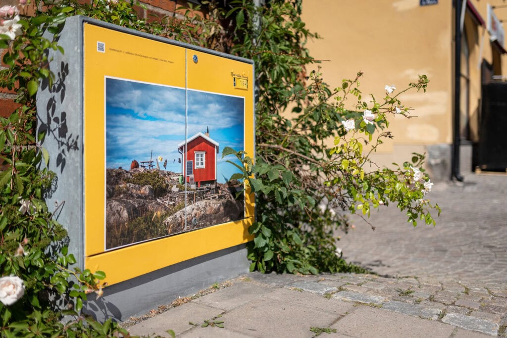 Bild på ett elskåp i centrala Norrtälje. Ett elskåp med konstmotiv av ett rött skärgårdshus, inramat av gul kant och omringat av rosor.