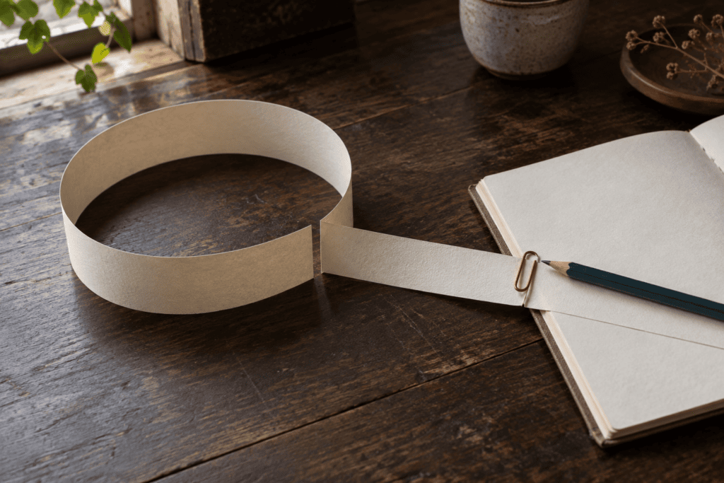 Composition de papier où une boucle s’ouvre vers un carnet, reliée par une bande et un trombone.