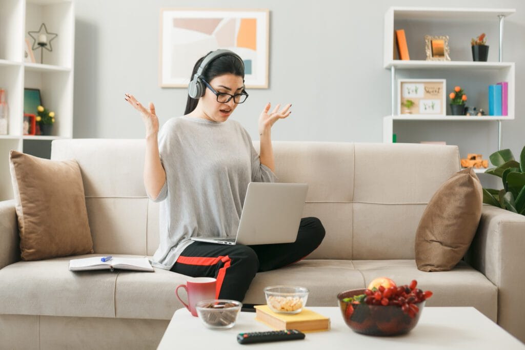 unpleased spreading hands young girl wearing glasses with headphones holding used laptop sitting sofa coffee table living room