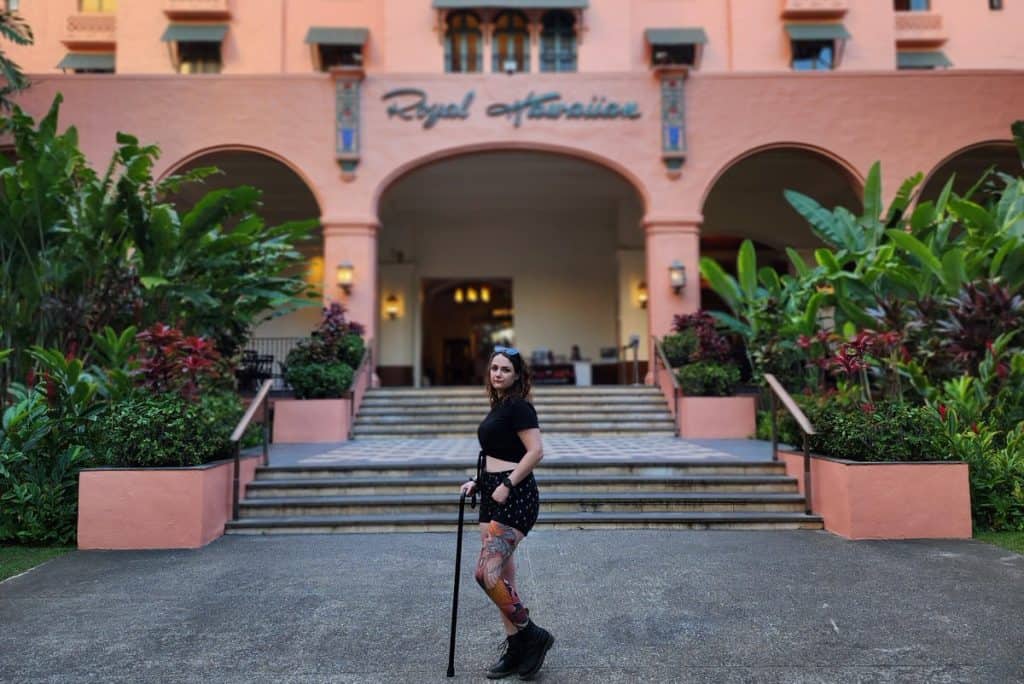 Phoenyx standing with a cane in front of the Royal Hawaiian Hotel’s iconic pink facade and grand staircase, surrounded by lush tropical greenery. The hotel’s arched entrance and sign are visible in the background.