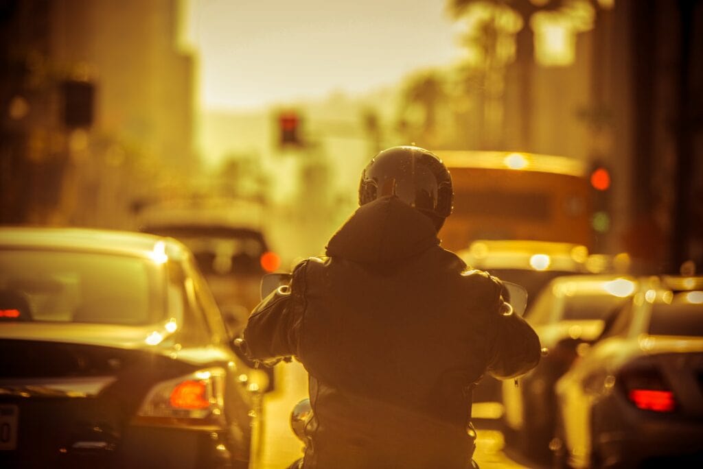 Motorcyclist seen from behind riding between lines of stopped cars in a city traffic jam at dusk