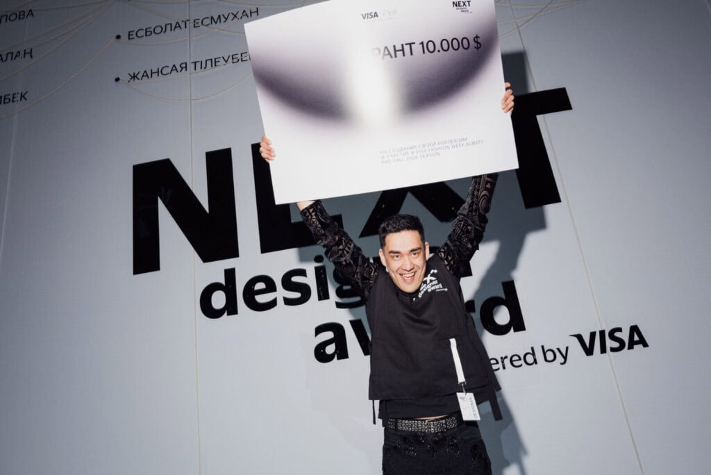 Designer Esbolat Esmukhanov holds up a $10,000 grant check at Visa Fashion Week Almaty after winning the NEXT Designer Award for his innovative "Fake Prada" collection.