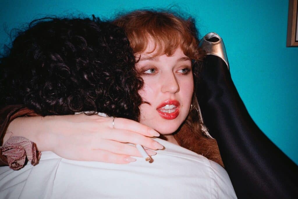 A red-haired woman with bold red lips embracing someone from behind against a turquoise wall, a cigarette between her fingers and a silver heel visible in the upper corner — photograph by Arthur Samier