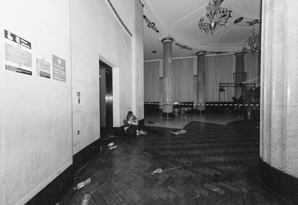 A person sits alone on the floor near an elevator in a dimly lit, empty lobby with high columns and a chandelier. Debris and cups are scattered on the parquet floor.