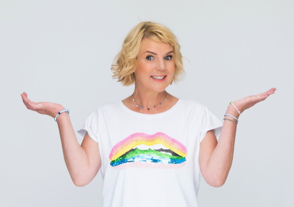 AIDA_Special_Comedy_Cruise_MirjaBoes__c_Lars_Laion - Michis Kreuzfahrten eine lächelnde person mit blonden lockigen haaren zeigt beide hände offen, gekleidet in einem weißen t-shirt mit buntem regenbogen-motiv auf einem neutralen hintergrund.