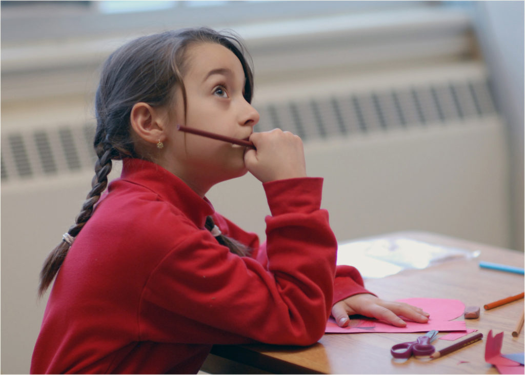 A little girl looking up thinking with paper, pencil crayons and scissors in front of her on the desk | Bishop Hamilton Montessori School in Ottawa