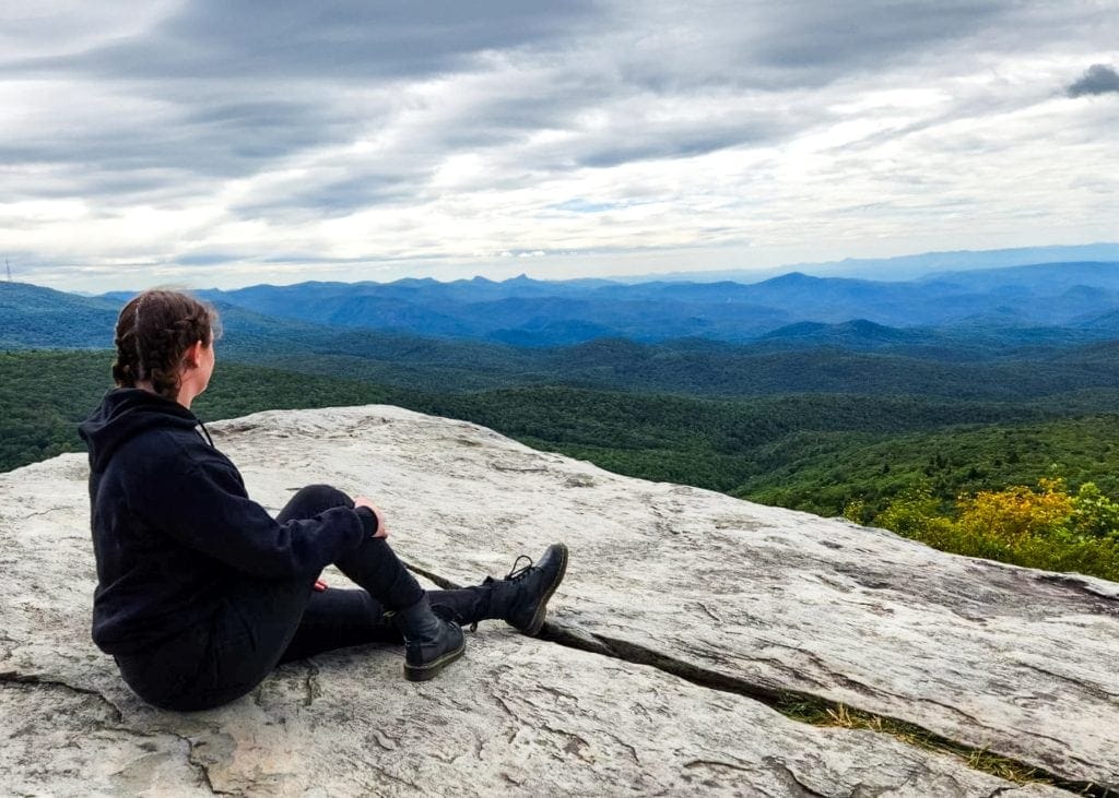 A woman in all black sitting on Rough Ridge rock overlooking the Blue Ridge Mountains on a cloudy day.
