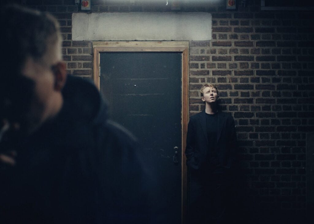 A person in dark clothing leans against a brick wall next to a door, looking up pensively. Another figure, blurred in the foreground, moves out of frame. The scene is dimly lit, creating a moody atmosphere.