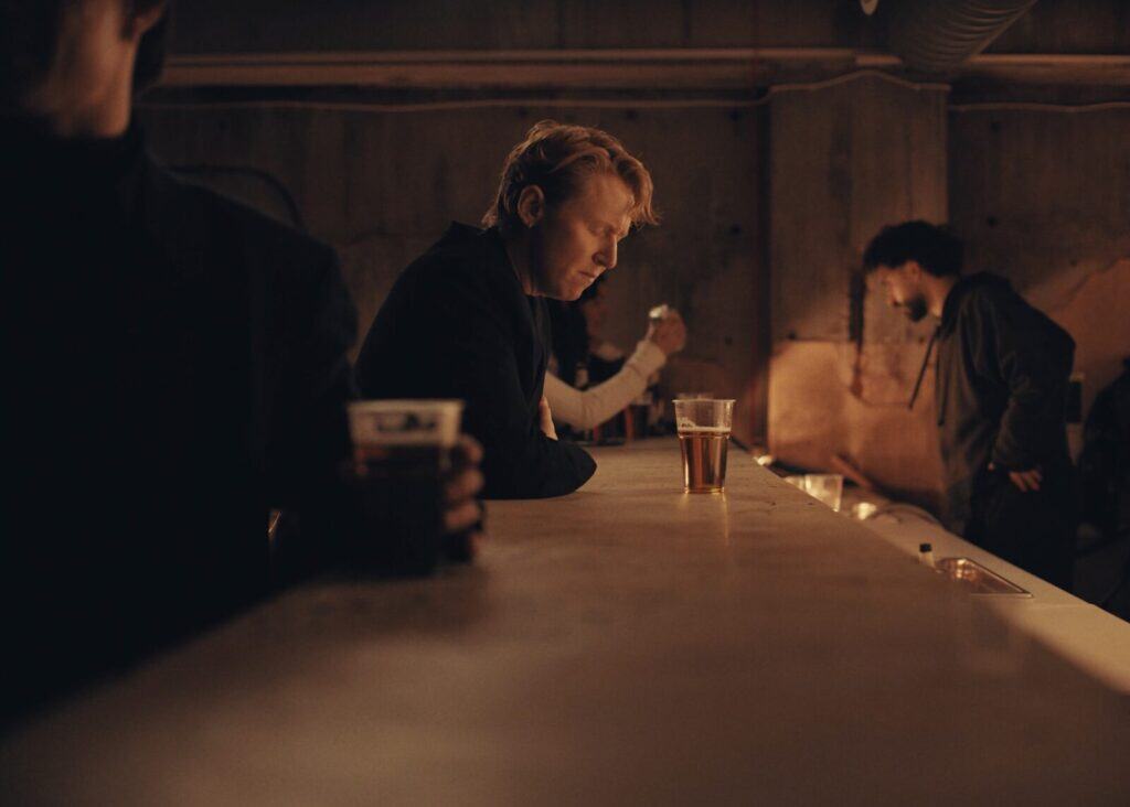 A man with light hair sits alone at a dimly lit bar, looking down thoughtfully near a glass of beer. Other people are in the background, one holding a drink, creating a moody and quiet atmosphere.