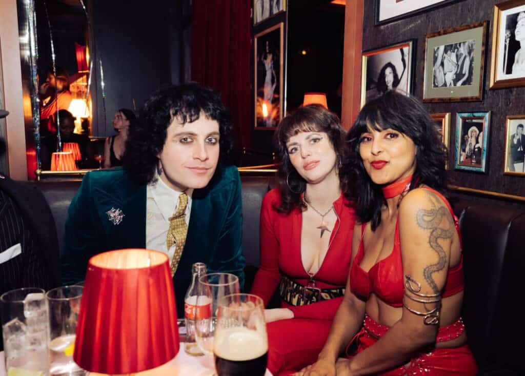 Three people in bold, retro-inspired outfits sit together in a dimly lit bar with red lamps and framed photographs on the walls, looking at the camera and smiling. Drinks are on the table in front of them at the Flamingo Club.