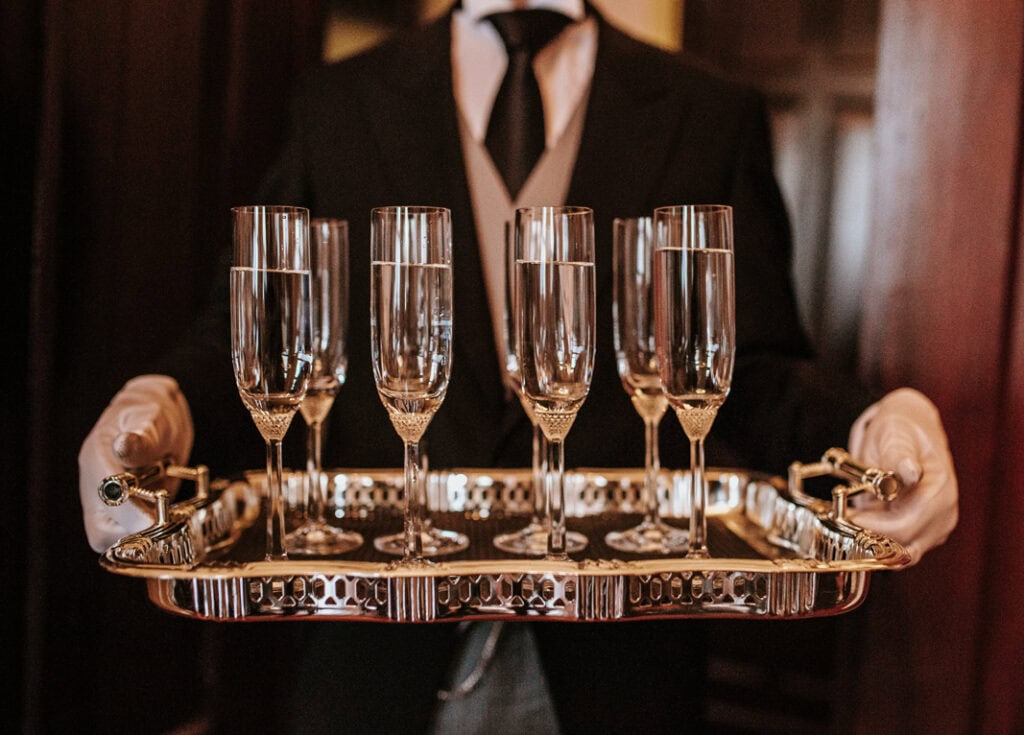A butler balances a tray with six sparkling glasses of champagne, highlighting a moment of luxury