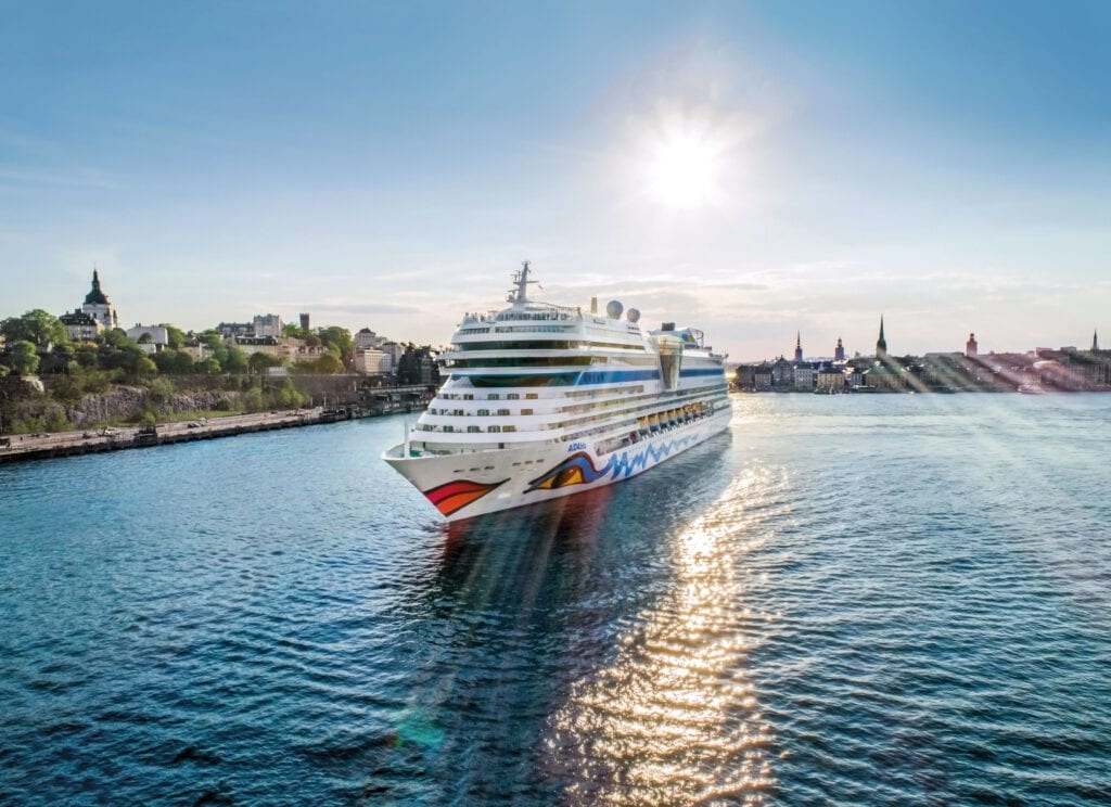 01_AIDAmar_c_FelixGaensicke - Michis Kreuzfahrten ein großes kreuzfahrtschiff fährt ruhig in einem hafen, umgeben von gebäuden und bäumen. die sonne scheint hell am himmel und reflektiert auf der wasseroberfläche.
