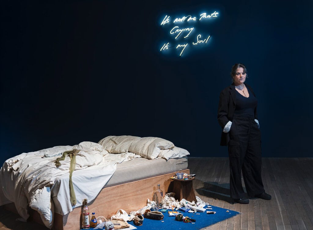 Artist Tracey Emin standing next to her famous installation My Bed at the Tate Modern, with a neon sign above her that reads "It's not me that's crying, it's my soul."