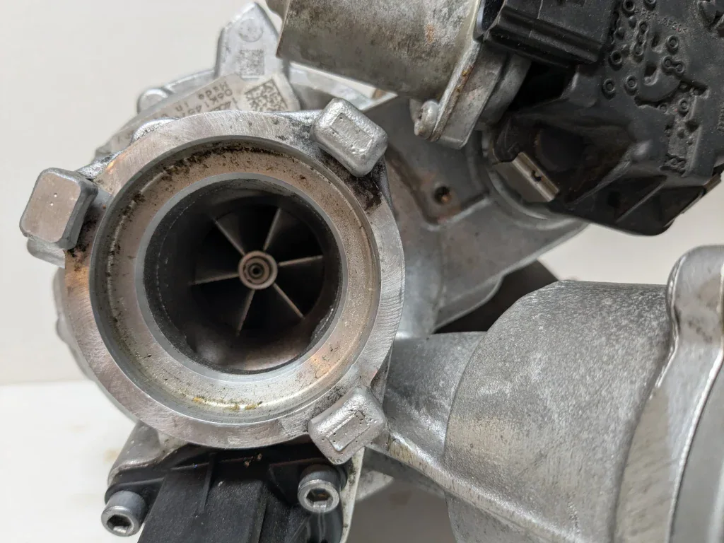 Detailed view of an IS20 turbocharger compressor side. The compressor wheel is visible inside the inlet, surrounded by a slightly dirty aluminum housing. A QR code sticker and electronic wastegate actuator are also visible. Useful for illustrating shaft-play inspection and visual checks.