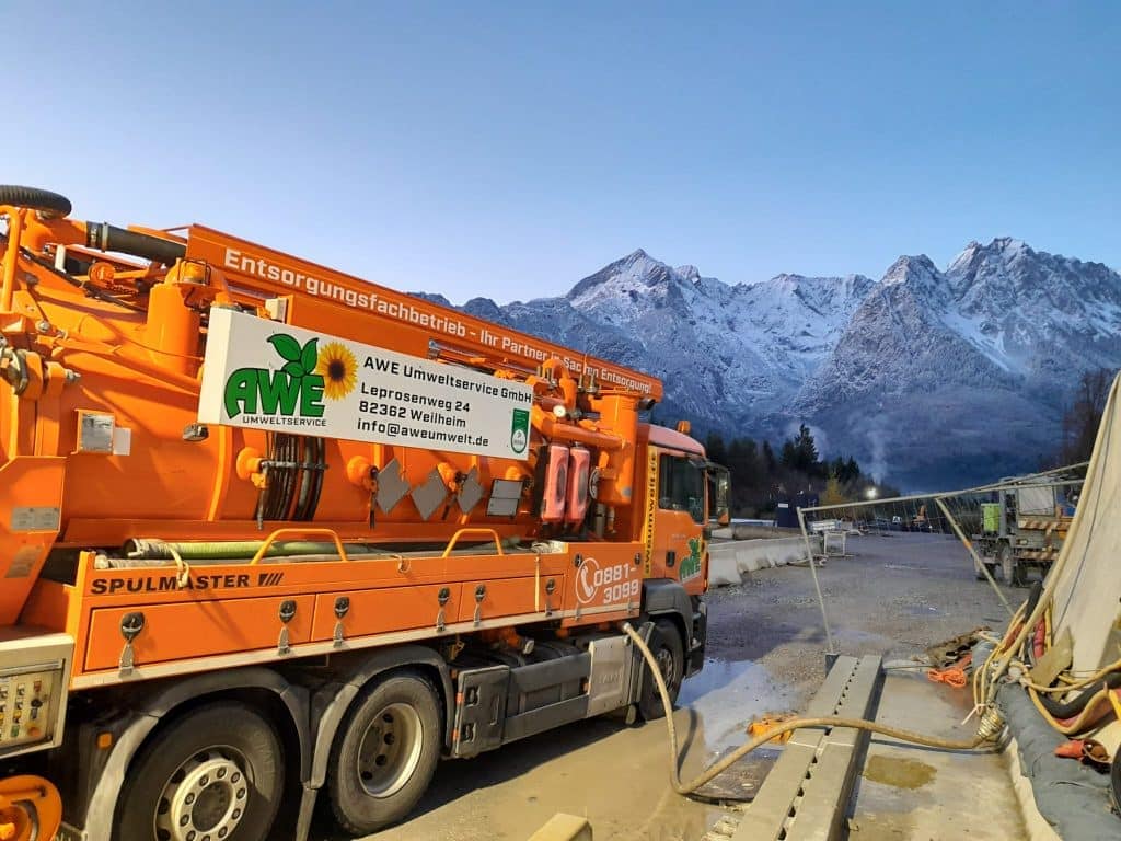 „Oranger Kanalspüler beim Wassertanken am frühen Morgen vor verschneiter Berglandschaft auf Baustelle“