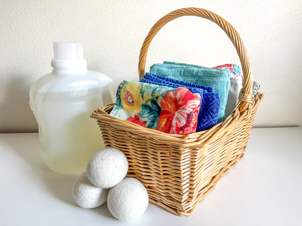 Basket of clean kitchen towels on a white dresser with natural laundry detergent and three wool dryer balls