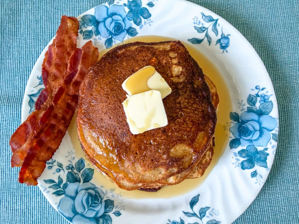 Landscape view of a stack of einkorn flour pancakes on a blue floral plate, served with butter, maple syrup, and two strips of bacon