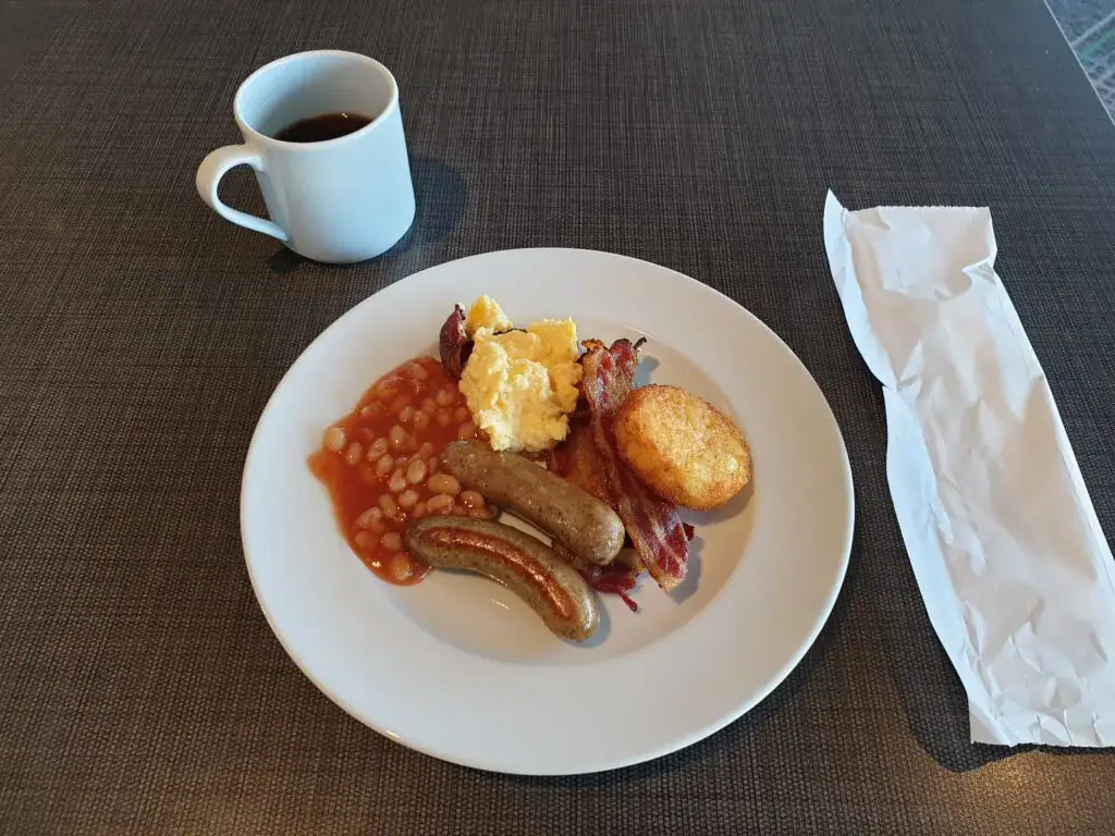 im vordergrund ein weißer teller mit einem english breakfast, würstl, speck, bohnen, kartoffelrösti und rührei. im hintergrund ein becher mit kaffee