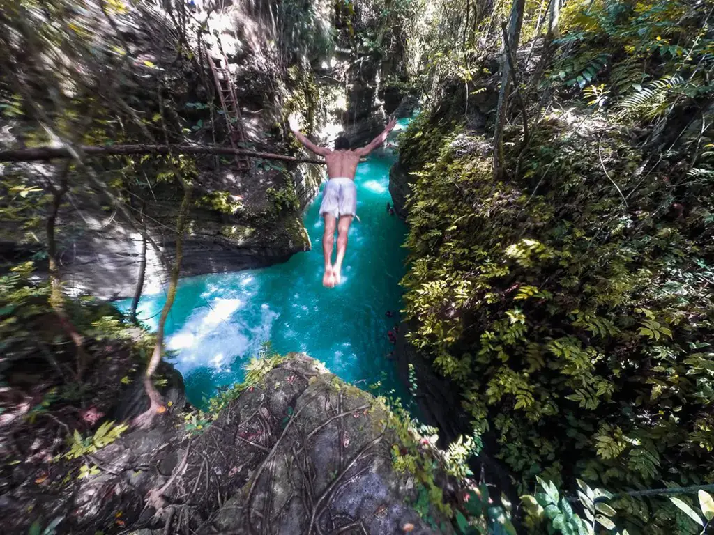 A picture of someone diving from a cliff into the water about 50 feet below at Kawasan Falls, in the Philippines.