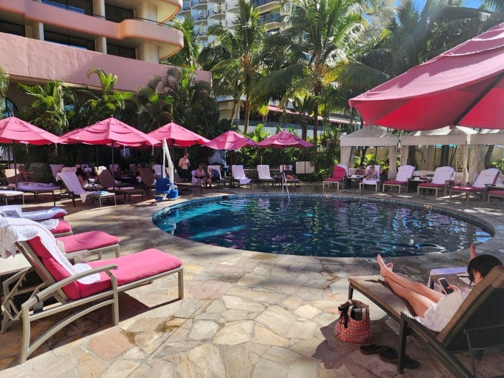 The Malulani Pool at The Royal Hawaiian Hotel in Waikīkī, Hawaii, surrounded by pink cushioned lounge chairs and bright pink umbrellas, with palm trees and cabanas adding tropical charm. Guests relax poolside in the sunshine near the hotel's signature pink architecture.