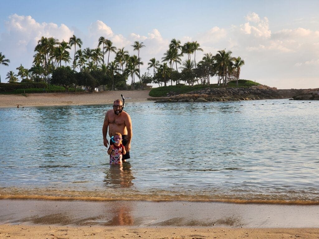 Justin and River enjoying the warm water at a serene beach in Kapolei, Oahu. Both are wearing snorkel gear, with lush palm trees and a sandy shoreline creating a tropical backdrop.