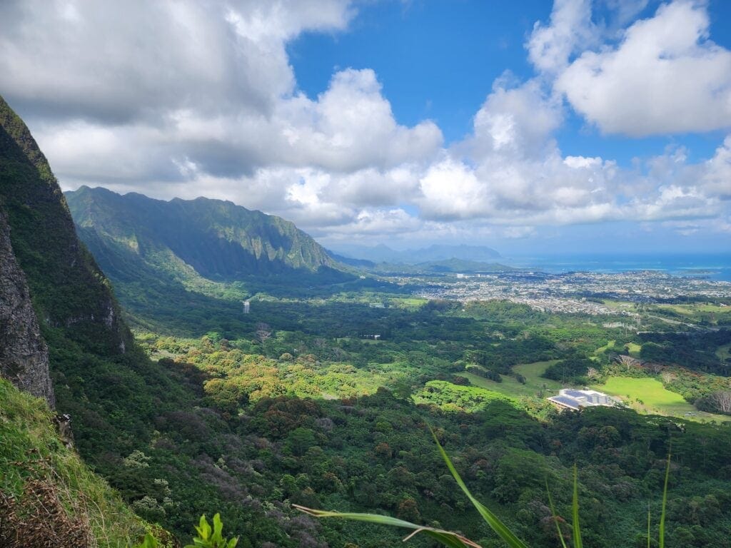 Breathtaking view from Pali Lookout on Oahu, Hawaii, showcasing lush green valleys, rugged mountains, and the sparkling blue Pacific Ocean under a partly cloudy sky.