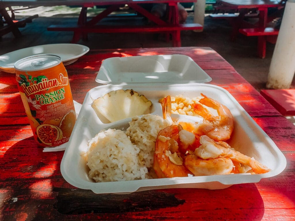 A takeout plate of garlic shrimp, white rice, corn, and a pineapple slice sits on a red picnic table next to a Hawaiian Sun Passion Orange juice can.