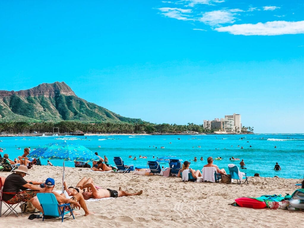 Crowds relaxing on the sandy shore of Waikīkī Beach with bright blue ocean waves and Diamond Head crater in the background under a sunny sky.