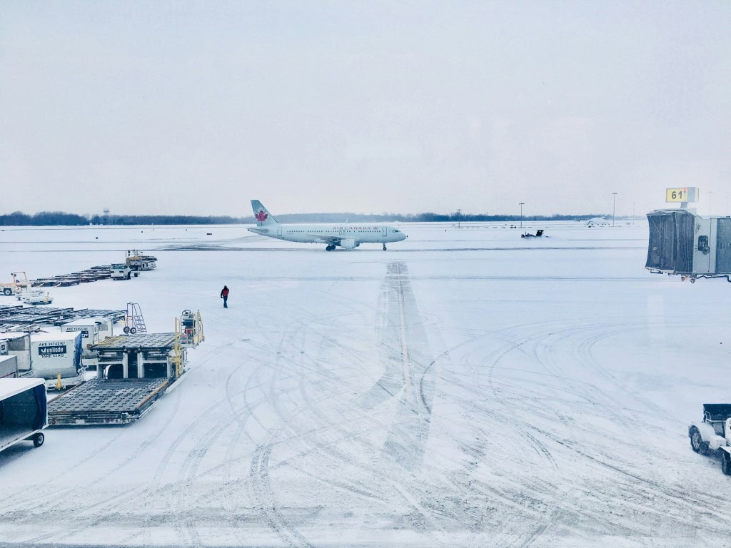 An airport scene covered in snow, featuring an Air Canada plane taxiing on a snowy runway with snow-covered ground services and baggage carts visible.