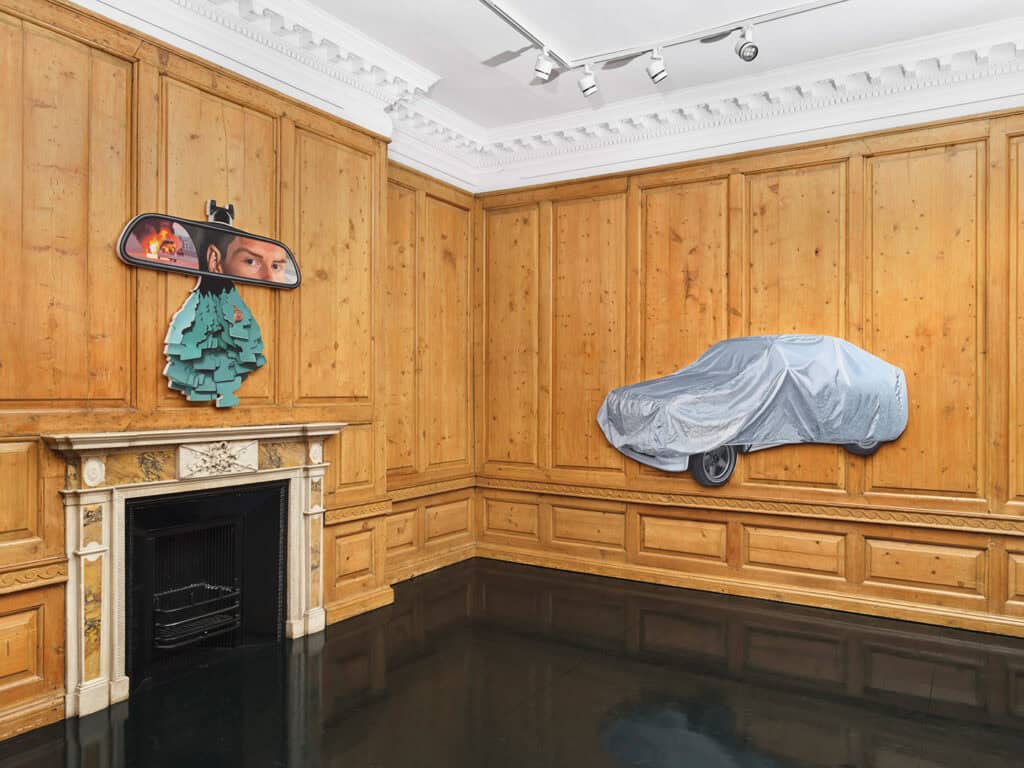 A wood-paneled room with a fireplace, featuring a large art piece above it resembling a car mirror with faces and air fresheners—part of the immersive Callum Eaton exhibition. A covered car is mounted on the wall, partially emerging from the paneling.