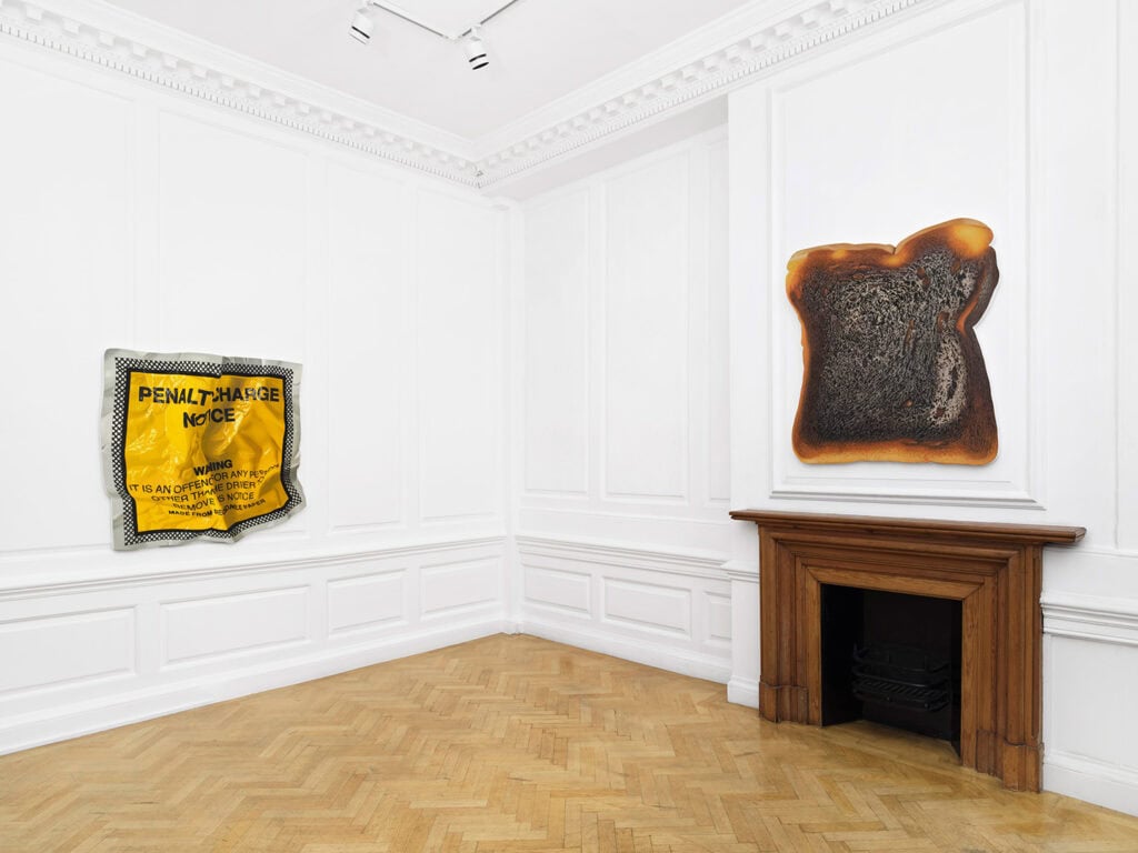 A white-walled gallery room with wood flooring features two artworks: a yellow penalty charge notice sign on one wall and a large, realistic sculpture of burnt toast above a wooden fireplace, exemplifying the playful spirit of the Callum Eaton exhibition.