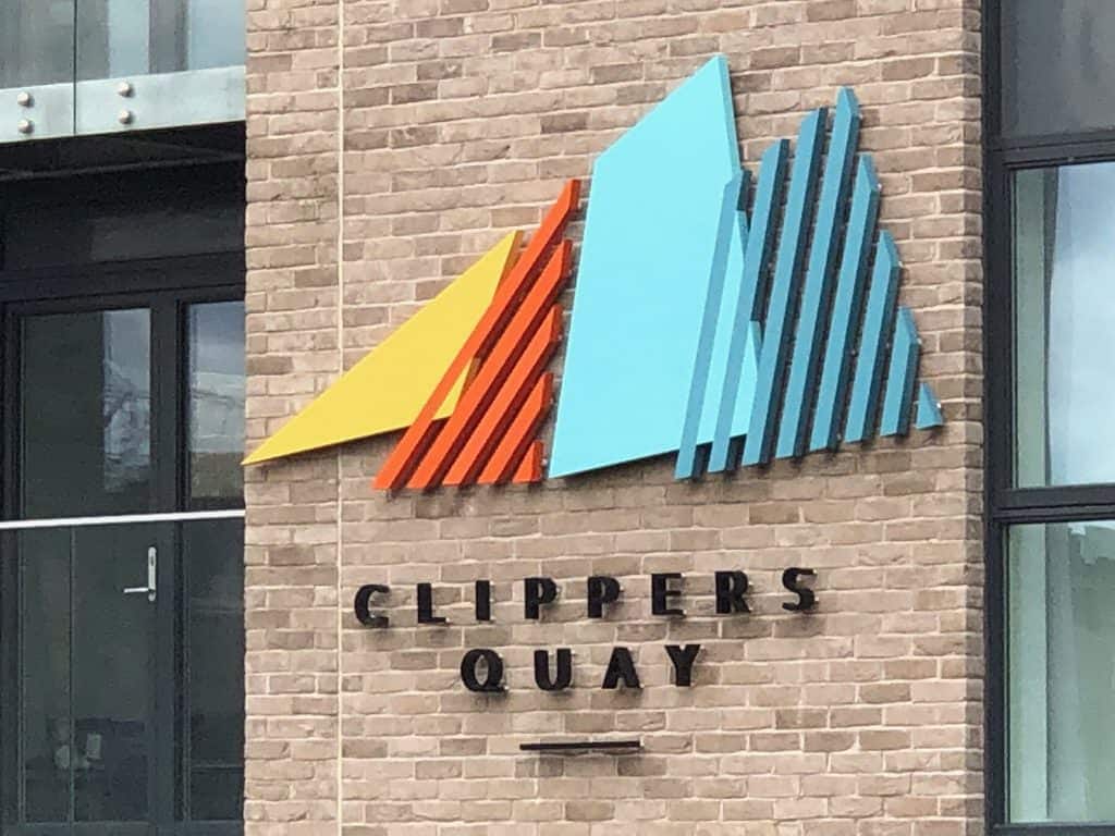 Multicoloured identity sign for Clippers Quay applied to a brick wall. The letters are black and applied as individual 3D characters. The logo type is composed of four overlapping sails – a yellow solid triangle, followed by an orange triangular shaped striated outline, a solid pale blue trapezoid and a further turquoise striated trapezoid.