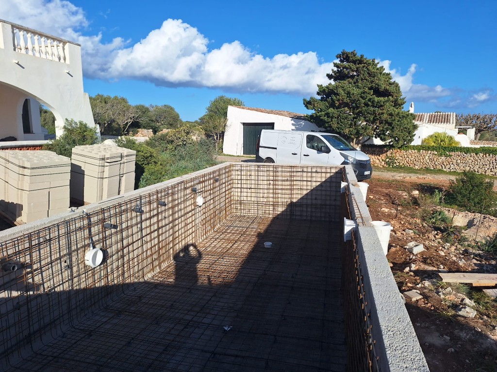 Construcción de piscina en Menorca, estructura con refuerzo de acero, encofrado y entorno rural con terraza blanca y coche blanco al fondo del terreno.