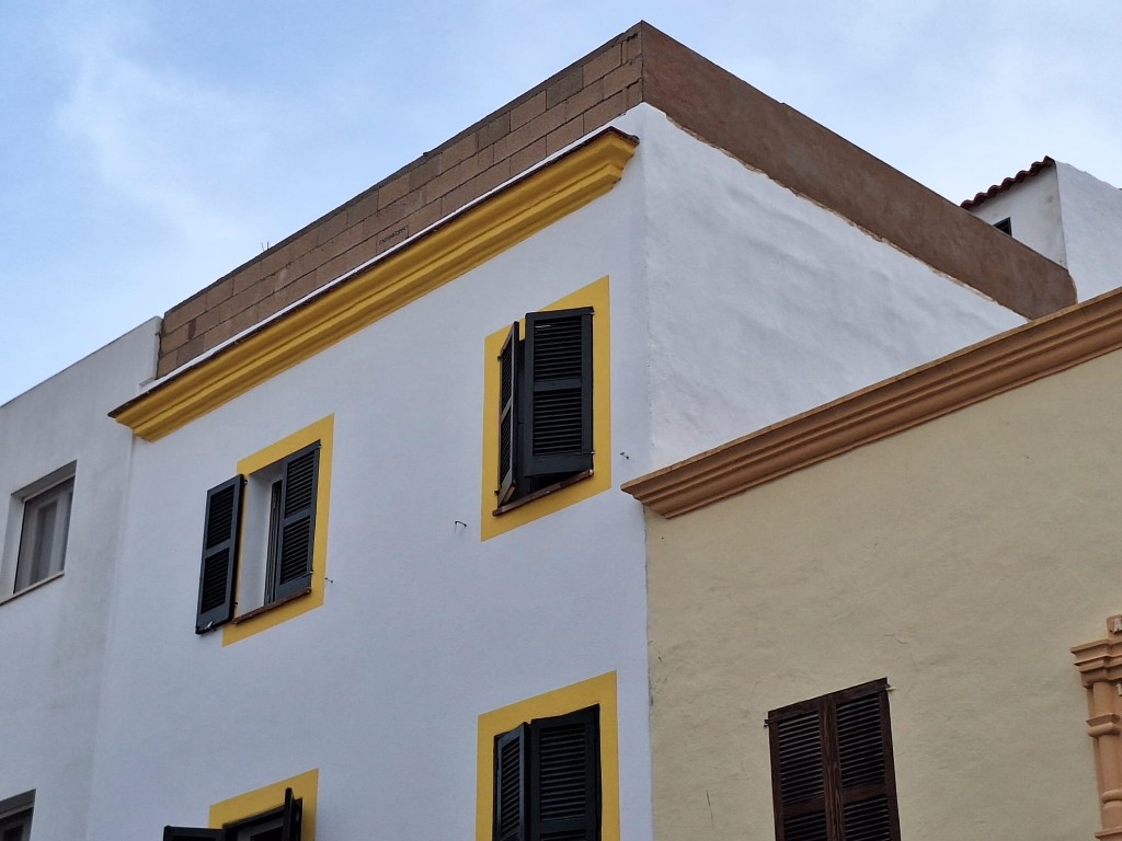 Fachada blanca con marcos y contornos amarillos y contraventanas negras en Menorca, detalle de vivienda mediterránea bajo cielo azul claro y luz suave.