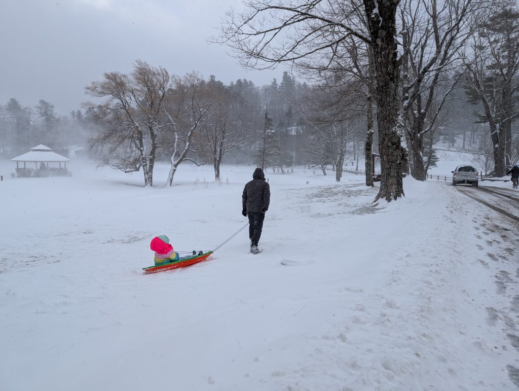 A man pulling a child on a sled through a snowy field at the Chetola Resort, with bare winter trees, a gazebo in the background, and a snow-covered hill ahead on a cold, overcast day.